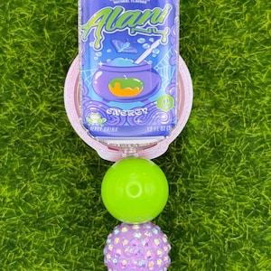 Puede incluir: Un carrete de insignia con un dise&ntilde;o de lata morada y azul, con el texto "Witch's Brew" y "Alani". El carrete tiene un dise&ntilde;o de cuentas verdes y moradas. El dise&ntilde;o de la lata incluye un caldero y las palabras "Energy Drink".