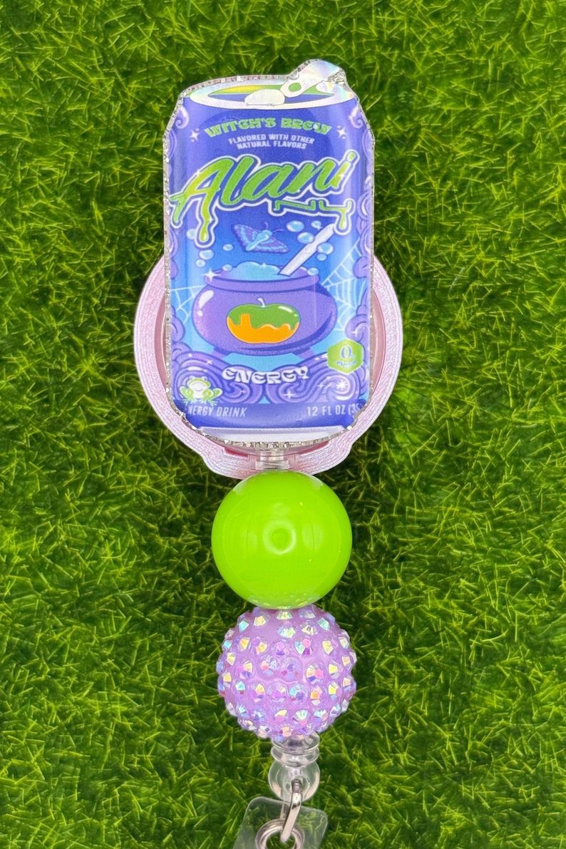 Puede incluir: Un carrete de insignia con un dise&ntilde;o de lata morada y azul, con el texto "Witch's Brew" y "Alani". El carrete tiene un dise&ntilde;o de cuentas verdes y moradas. El dise&ntilde;o de la lata incluye un caldero y las palabras "Energy Drink".