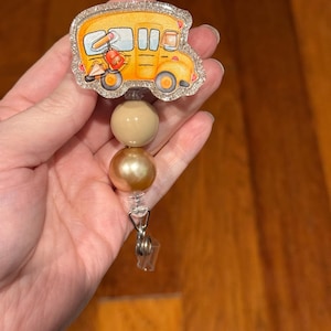 May include: A yellow school bus with a glitter finish is attached to a clear badge reel with two beige and gold beads.