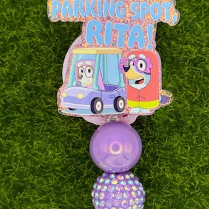 May include: A purple and blue badge reel with the text "NICE PARKING SPOT, RITA!". The badge reel features cartoon characters in a car and a purple beaded design.