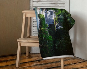 May include: A green and white throw blanket with a photo of a waterfall and lush greenery. The blanket is draped over a wooden stool.