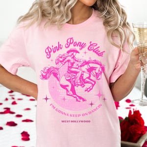 May include: Pink t-shirt with a graphic of a woman riding a horse in front of a disco ball. The text "Pink Pony Club" is above the graphic and "I'm gonna keep on dancin' West Hollywood" is below the graphic.