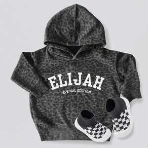 Pode incluir: Camisola de capuz preta e branca com estampa de leopardo e o texto "ELIJAH SPECIAL EDITION" em branco. A camisola é combinada com meias xadrez pretas e brancas.