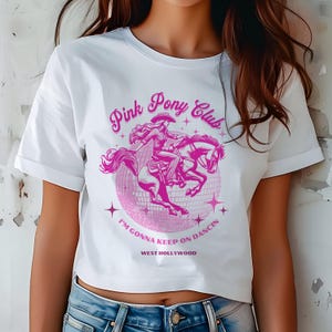 May include: White cropped t-shirt with a pink graphic of a woman riding a horse in front of a disco ball. The graphic says "Pink Pony Club" and "I'm gonna keep on dancin' West Hollywood".