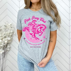 May include: A gray t-shirt with a pink graphic of a woman riding a horse. The graphic says "Pink Pony Club" and "I'm gonna keep on dancin' West Hollywood".