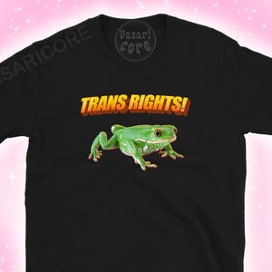 Puede incluir: Camiseta negra con una rana verde y el texto "TRANS RIGHTS!" en amarillo y rojo. La rana tiene sombreado realista. La camiseta tiene mangas cortas y cuello redondo.
