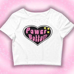 Puede incluir: Top corto blanco con un gráfico de corazón rosa que dice "Power Bottom" con tres estrellas amarillas.