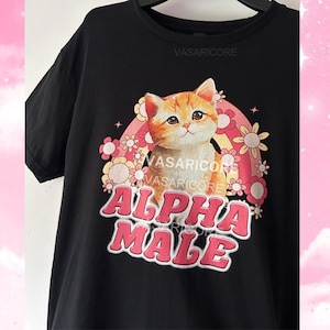 Puede incluir: Camiseta negra con un gráfico de un gato frente a un arcoíris y flores. El texto "ALPHA MALE" está impreso en letras rosas.