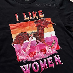 K&ouml;nnte beinhalten: Schwarzes T-Shirt mit einem roten Drachen, der Feuer spuckt, vor einem gestreiften Hintergrund in Pink, Orange und Gelb. Der Text "I LIKE WOMEN" ist in Pink unter dem Drachen aufgedruckt.