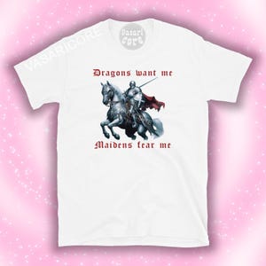 Może przedstawiać: Biała koszulka z nadrukiem przedstawiającym rycerza na białym koniu. Tekst "Dragons want me" i "Maidens fear me" jest nadrukowany na koszulce na czerwono.