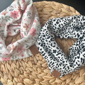 Könnte beinhalten: Zwei Stoff-Halstücher. Eines ist weiß mit rosa und pfirsichfarbenen Blumenmustern. Das andere ist schwarz-weiß mit Leopardenmuster. Beide haben ein braunes Lederetikett mit einem Logo. Sie werden auf einer gewebten Matte präsentiert.