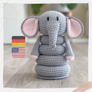 Puede incluir: Un juguete de elefante de crochet gris con orejas rosas. El elefante es un juguete apilable con cuatro anillos. Los anillos también son grises y de crochet.