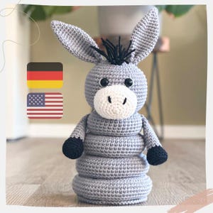 以下が含まれることがあります： 黒い目と黒いたてがみを持つグレーの編みぐるみロバの玩具。ロバは4つのリングが付いた積み重ね式玩具です。