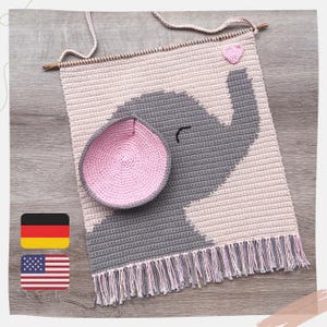 Pode incluir: Um pendurador de parede de crochê feito à mão com um desenho de elefante cinza sobre um fundo rosa e cinza. O elefante tem uma orelha rosa e um pequeno coração rosa. O pendurador de parede é finalizado com franjas e uma vara de madeira para pendurar.