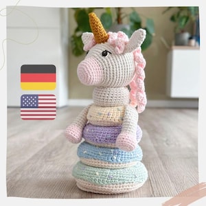 Può includere: Un giocattolo di unicorno all'uncinetto con una criniera e un corno rosa. L'unicorno è seduto su una pila di anelli colorati all'uncinetto.