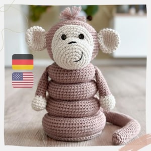 Peut inclure: Un jouet singe en crochet avec une pile d'anneaux pour le corps. Le singe est rose avec des traits du visage blancs et une longue queue.