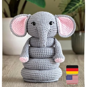 Könnte beinhalten: Ein graues, gehäkeltes Elefanten-Spielzeug aus gestapelten Ringen. Der Elefant hat große Ohren mit rosa Innenseiten, schwarze Sicherheitsaugen, einen Rüssel und kleine rosa Akzente an den Füßen. Eine deutsche Flagge und eine US-Flagge sind abgebildet.