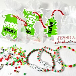 Peut inclure: Sacs cadeaux de Noël avec des étiquettes vertes représentant un elfe, un élan et un sapin, chacune avec un nom. Des bracelets de perles avec des noms sont présents, ainsi que des perles en vrac. Une carte de nom indique "JESSICA".