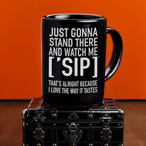 May include: Black ceramic mug with white text that reads "Just gonna stand there and watch me ['SIP] That's alright because I love the way it tastes". The mug is sitting on top of a black vintage suitcase.