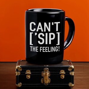 May include: Black ceramic mug with white text that reads "Can't 'Sip' the Feeling!" The mug is sitting on top of a black leather suitcase with gold accents.