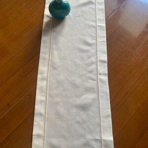 May include: White linen table runner with white embroidered edging. The runner is on a wooden table with a teal blue vase with sticks in it.