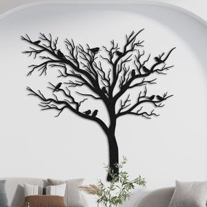 May include: Black metal tree wall art with birds perched on the branches. The tree is mounted on a white wall above a modern living room set. The tree's silhouette is detailed, creating a striking contrast against the light background.