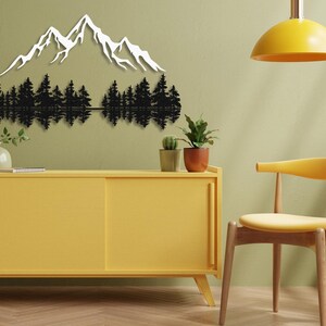 May include: Black metal wall art depicting a mountain range with trees reflected in a lake. The art is mounted on a light green wall above a yellow cabinet and a yellow chair.