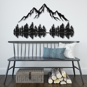 May include: Black metal wall art depicting a mountain range with a reflection of the mountains in a lake below. The mountains are silhouetted against a white background. The art is displayed on a white wall above a gray bench with pillows.