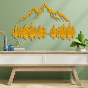 May include: A gold metal wall art sculpture depicting a mountain range with trees reflected in a lake. The sculpture is mounted on a light green wall above a light wood console table.