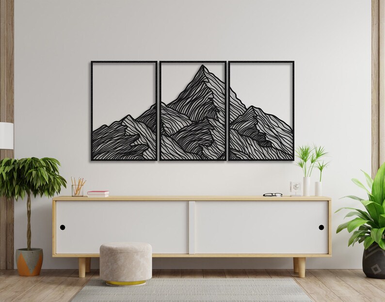 May include: A black metal wall art sculpture depicting a mountain range with a minimalist line art design. The sculpture is made up of three separate pieces, each depicting a different section of the mountain range. The sculpture is mounted on a white wall above a wooden cabinet.