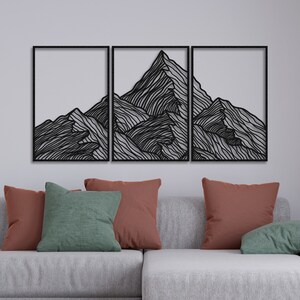 May include: A black metal wall art sculpture depicting a mountain range with a minimalist line art design. The sculpture is divided into three sections and is mounted on a white wall above a gray couch with pillows.
