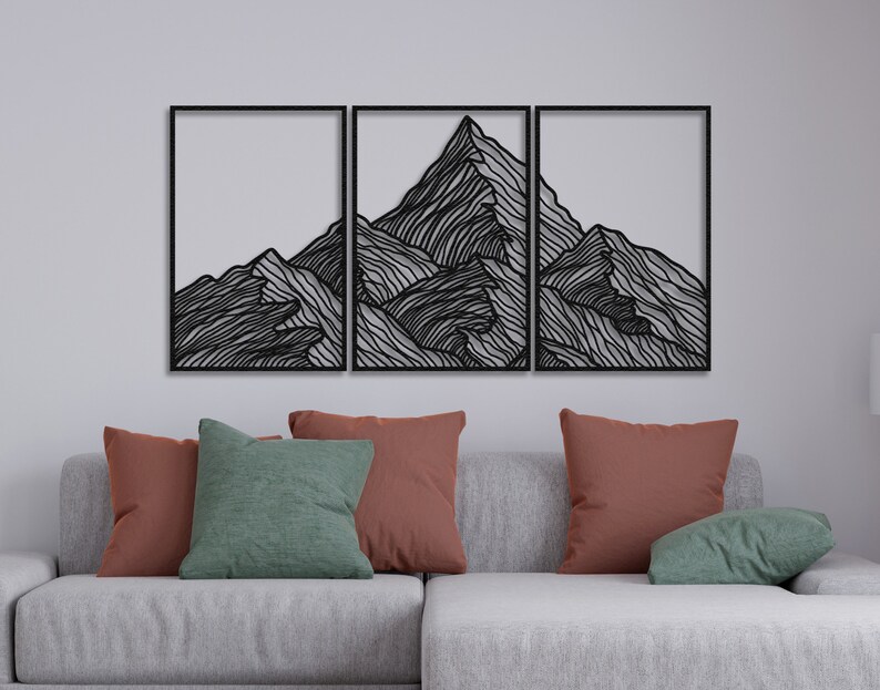 May include: A black metal wall art sculpture depicting a mountain range with a minimalist line art design. The sculpture is divided into three sections and is mounted on a white wall above a gray couch with pillows.