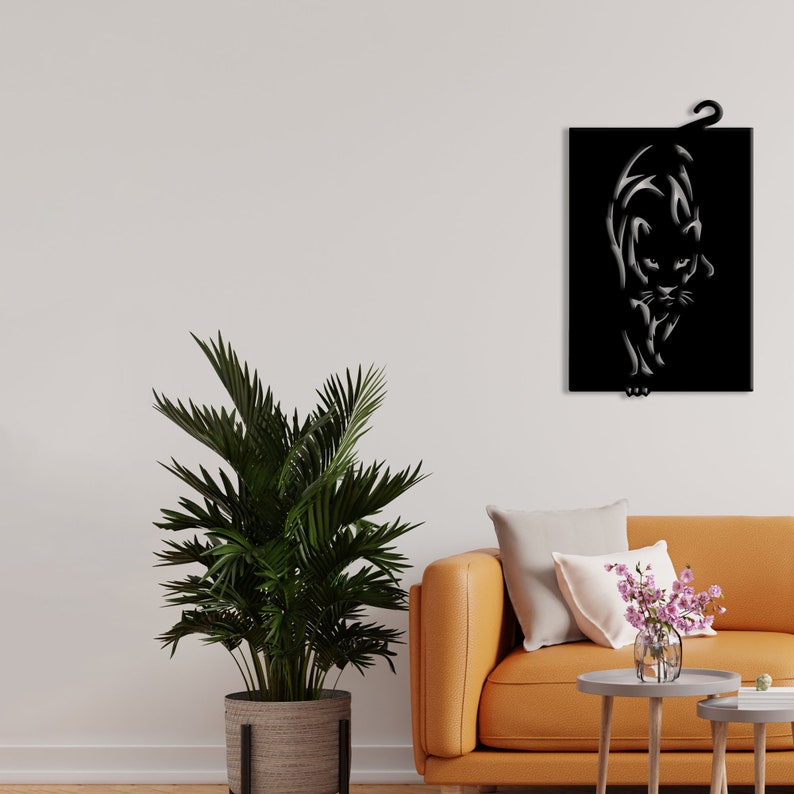 K&ouml;nnte beinhalten: Schwarze Wandkunst aus Metall mit einer stilisierten Silhouette eines Panthers. Der Panther blickt nach vorne, der Kopf leicht geneigt. Die Kunst h&auml;ngt an einer wei&szlig;en Wand &uuml;ber einem braunen Sofa mit einem wei&szlig;en Kissen und einer Vase mit rosa Blumen auf einem kleinen Tisch.