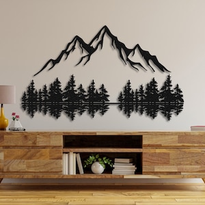 May include: Black metal wall art depicting a mountain range with a reflection of trees in a lake. The art is mounted on a light brown wooden wall shelf.