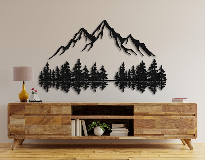 May include: Black metal wall art depicting a mountain range with a reflection of trees in a lake. The art is mounted on a light brown wooden wall shelf.