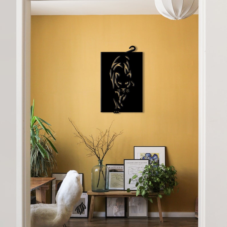 K&ouml;nnte beinhalten: Schwarze Wandkunst aus Metall mit einer Silhouette einer Katze, deren Kopf zur Seite gedreht ist. Die Katze blickt auf den Betrachter und hat einen langen Schwanz. Die Kunst ist an einer gelben Wand in einem Raum mit einem Tisch und St&uuml;hlen angebracht.