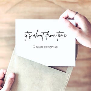 May include: A white rectangular greeting card featuring the black text "it's about damn time" in a cursive script font, followed by the text "I mean congrats" in a typewriter-style font. The card is partially inserted into a light brown envelope.