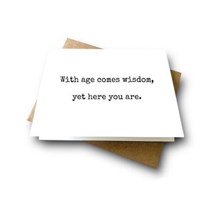 May include: A white rectangular greeting card with black typewriter-style text that reads, With age comes wisdom, yet here you are. The card is positioned on top of a brown kraft paper envelope, creating a simple and clean visual presentation.