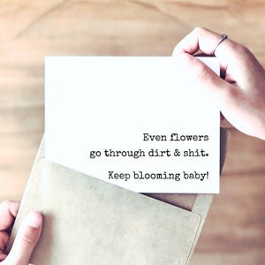 May include: A white rectangular greeting card partially inserted into a light brown envelope. The card features black typewriter-style text that reads: Even flowers go through dirt & shit. Keep blooming baby! The card is held against a wooden surface.