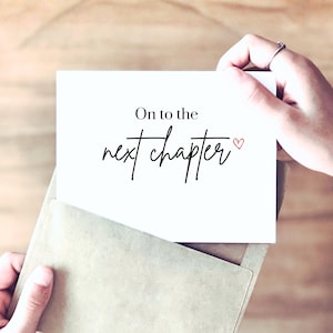 May include: A white rectangular greeting card featuring the black printed text On to the next chapter with a small red heart symbol at the end. The card is partially inserted into a light brown paper envelope, showcasing a clean and simple design.