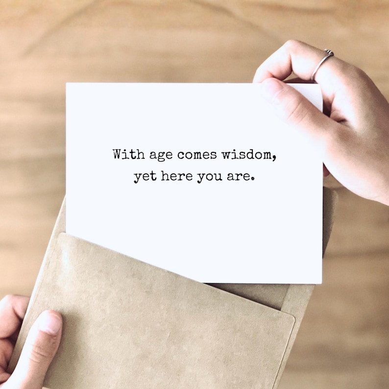 May include: A white rectangular greeting card featuring the black printed text "With age comes wisdom, yet here you are." The card is partially inserted into a light brown kraft paper envelope, showcasing a clean and simple design.