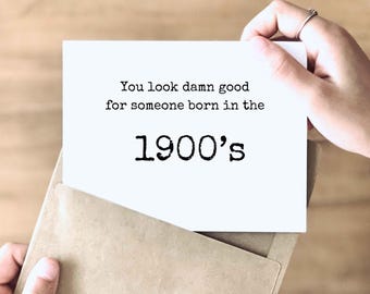 Może przedstawiać: Biała prostokątna kartka okolicznościowa z czarnym tekstem w stylu maszyny do pisania o treści: You look damn good for someone born in the 1900's. Kartka jest częściowo włożona do jasnobrązowej papierowej koperty i ma prosty, przejrzysty wygląd.