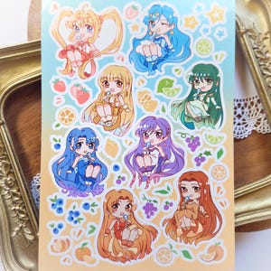 Può includere: Un foglio di adesivi con illustrazioni in stile anime di ragazze con capelli e abiti colorati, ognuna con un lecca-lecca. Il foglio include illustrazioni di frutta come fragole, limoni e uva. Lo sfondo è un gradiente giallo e blu.