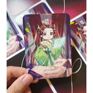 Puede incluir: Una pegatina rectangular con un personaje chibi con un atuendo verde y dorado, sosteniendo un abanico y un tablero de juego. El personaje tiene el pelo largo y castaño sobre un fondo rojo oscuro con confeti colorido.