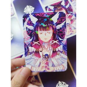 Puede incluir: Una tarjeta rectangular con una ilustración de estilo anime de un personaje con cabello largo y oscuro, alas blancas y un vestido blanco. El personaje tiene los ojos cerrados y sostiene un objeto pequeño. La tarjeta tiene un fondo morado y azul.