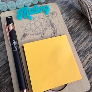 May include: A wooden notepad with a sea turtle design and the name "Mallory" in turquoise. A black pen with rose gold accents and the name "Mallory" is next to a yellow sticky note pad. Decorative beads are in the background.