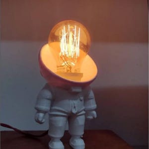 Puede incluir: Lámpara blanca con forma de astronauta y una gran bombilla como cabeza. La bombilla emite un brillo cálido y anaranjado, y la figura del astronauta está detallada con un diseño de traje espacial. La lámpara está enchufada con un cable negro.