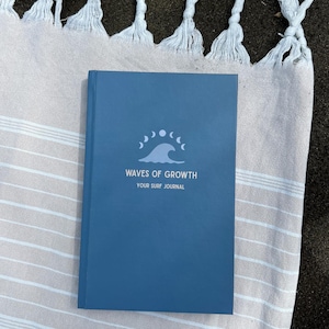 Könnte beinhalten: Ein blaues Notizbuch mit dem Text "WAVES OF GROWTH YOUR SURF JOURNAL" und einer Wellengrafik. Das Notizbuch liegt auf einem gestreiften beige-weißen Handtuch mit Fransen.