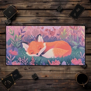 May include: A rectangular desk mat featuring a sleeping fox illustration. The fox is orange and white, nestled in a field of green grass and pink flowers. The background is a soft pink and purple. The mat is surrounded by wooden dice, cards, and a small cup.