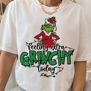 Op de afbeelding: Wit T-shirt met een Grinch-personage in rood en groen, versierd met kerstverlichting. De tekst "Feeling extra GRINCHY today" staat eronder gedrukt, perfect voor de feestdagen.
